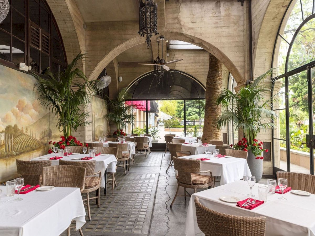 An elegant indoor courtyard dining area with set tables, red napkins, palms, and a grand arched ceiling with a chandelier.