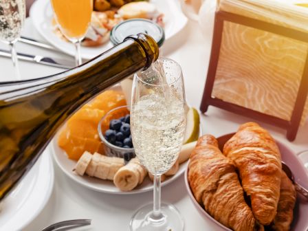 A sparkling drink is being poured from a bottle into a champagne flute on a breakfast tray with pastries and fruit, for a brunch vibe.