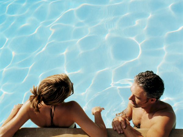 Two people relax poolside on a sunny day as clear blue water glitters with ripples.