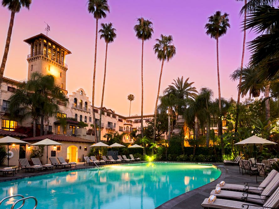 A resort courtyard with a glowing outdoor pool, lounge chairs, and tall palm trees at sunset.