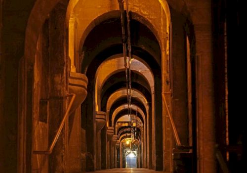 A long, dim industrial tunnel with repeating arched openings, metal pipes, and warm glowing lights along the corridor.
