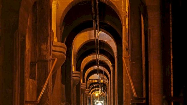 A long, dim industrial tunnel with repeating arched openings, metal pipes, and warm glowing lights along the corridor.