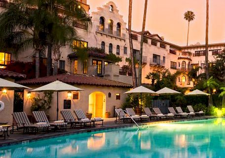 A hotel courtyard with a glowing swimming pool, palm trees, and warm-lit buildings at sunset.