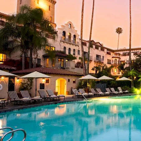 A hotel courtyard with a glowing swimming pool, palm trees, and warm-lit buildings at sunset.