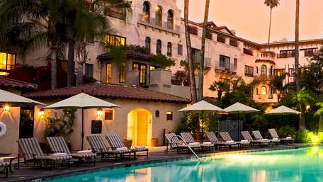 A hotel courtyard with a glowing swimming pool, palm trees, and warm-lit buildings at sunset.