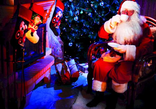 Santa in a red suit sits by a glowing fireplace, sipping a drink, with a festive tree and hanging ornaments behind him.