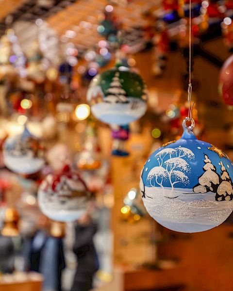 A close-up of hanging Christmas baubles, with a warm, festive, blurred background of ornaments and lights behind them.